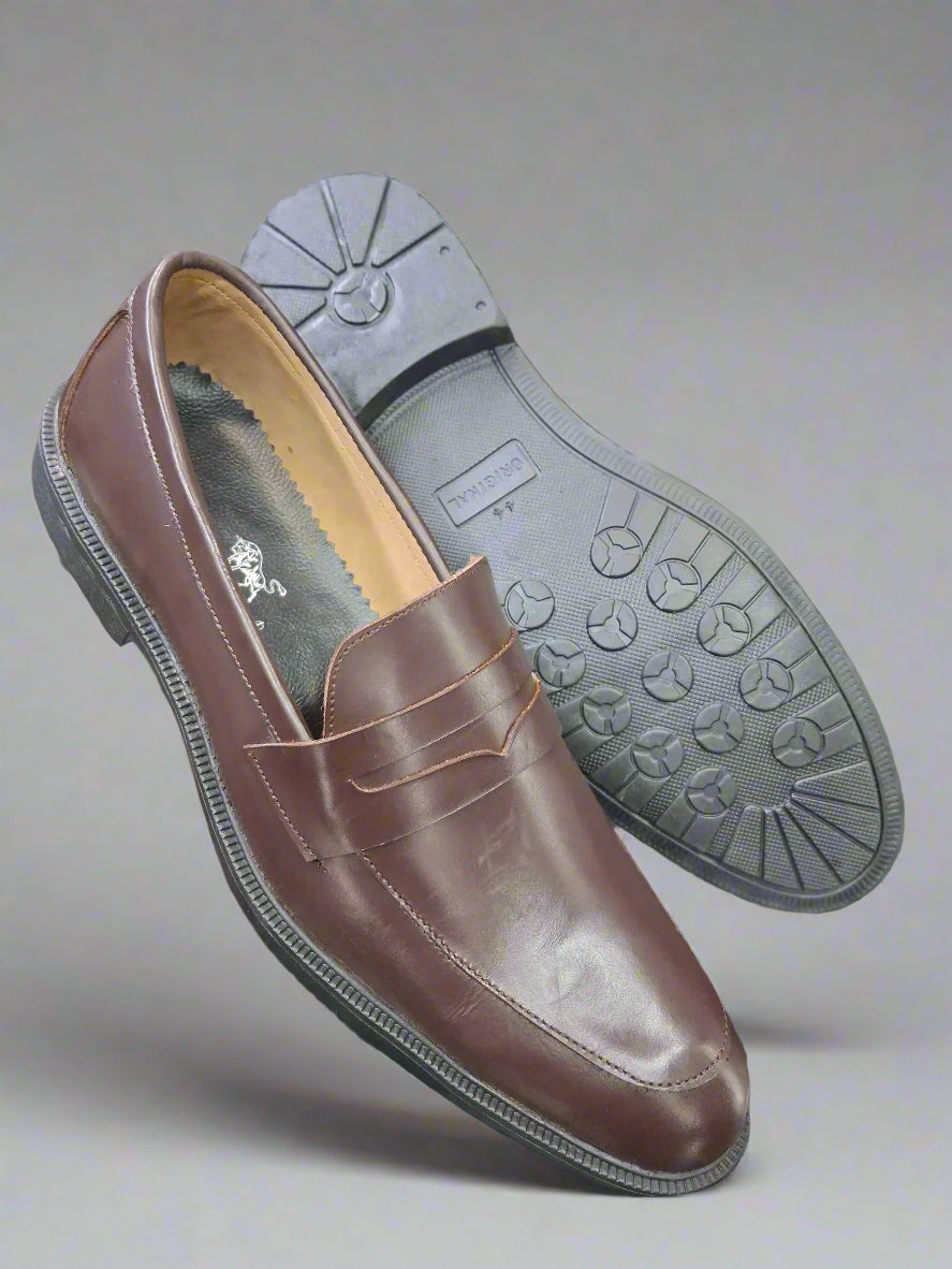 Brown loafer shoe with gray sole held by a person against a blurred background of shoes.