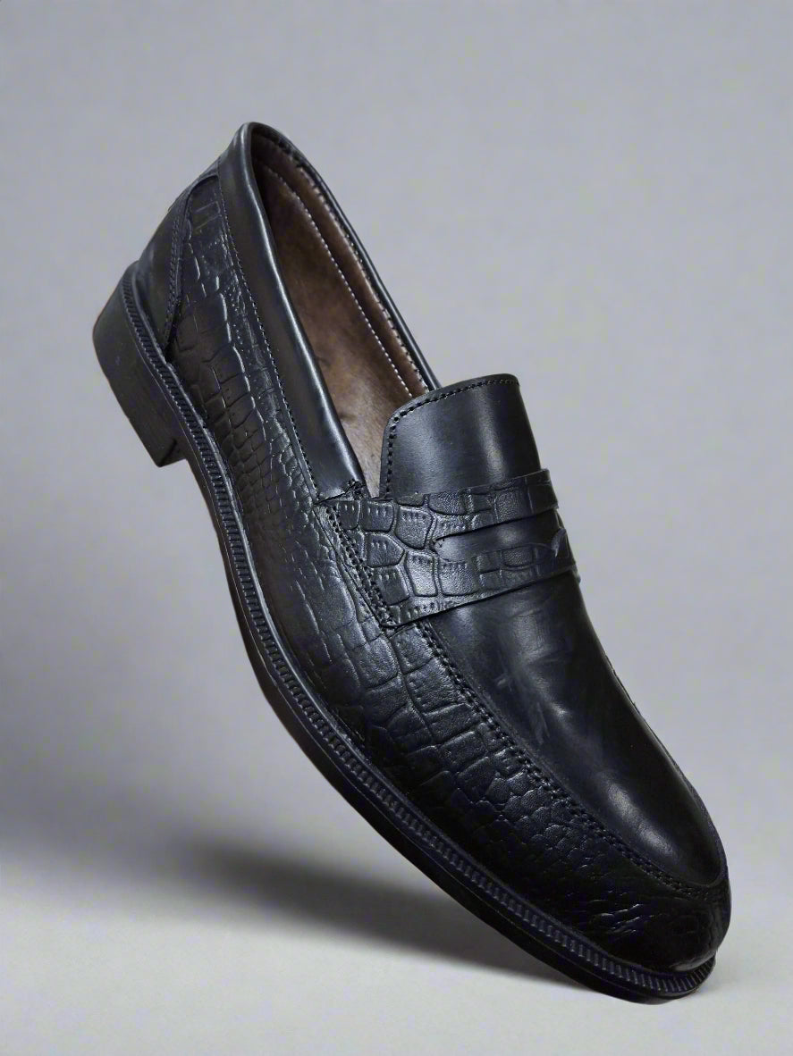Black leather loafer shoe held by a hand on a light-colored floor.