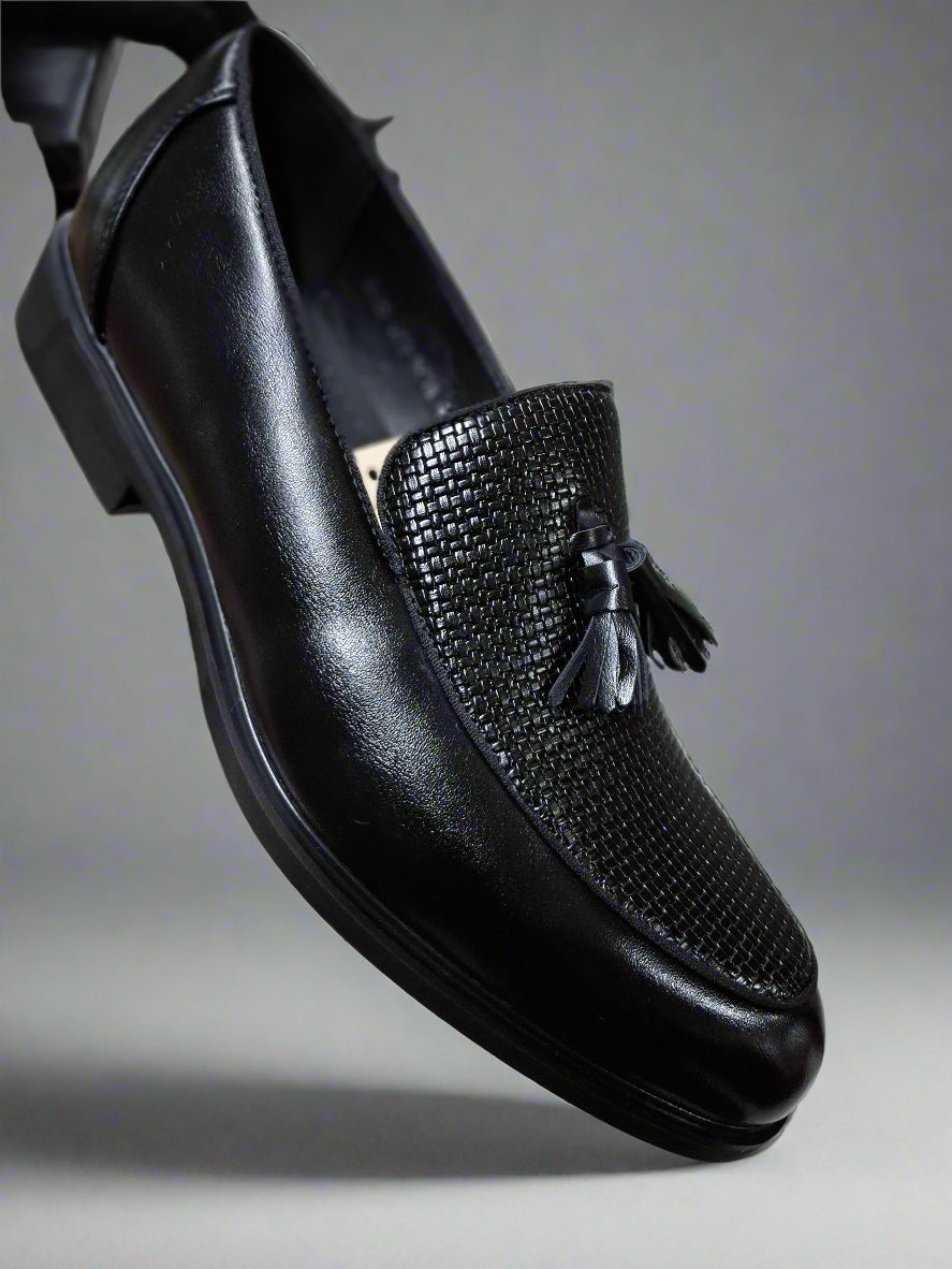 Black leather loafer with tassel held by a hand on a tiled floor.