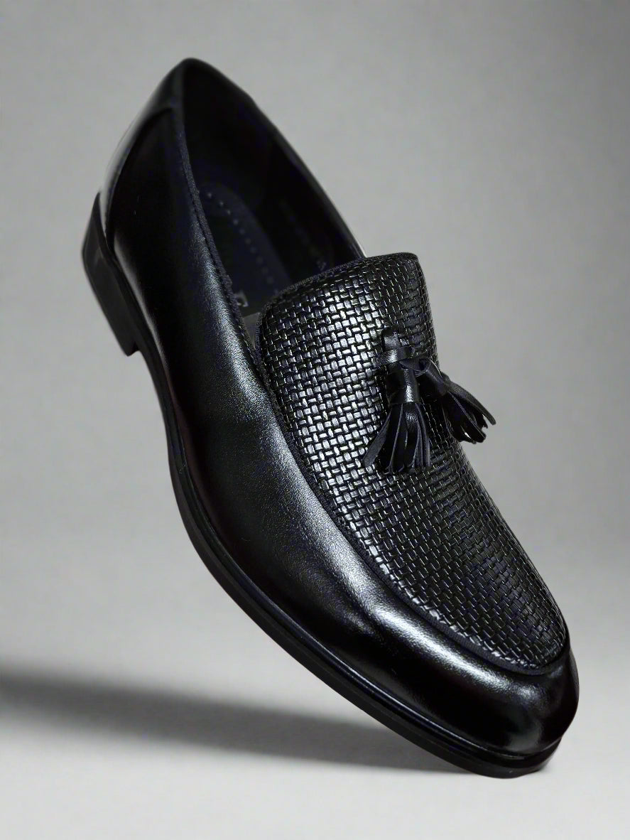 Black loafer shoe with tassel held by a hand against a neutral background