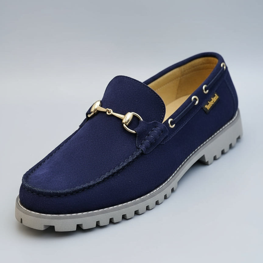 Blue suede loafer with gold buckle held by a hand, blurred store background.