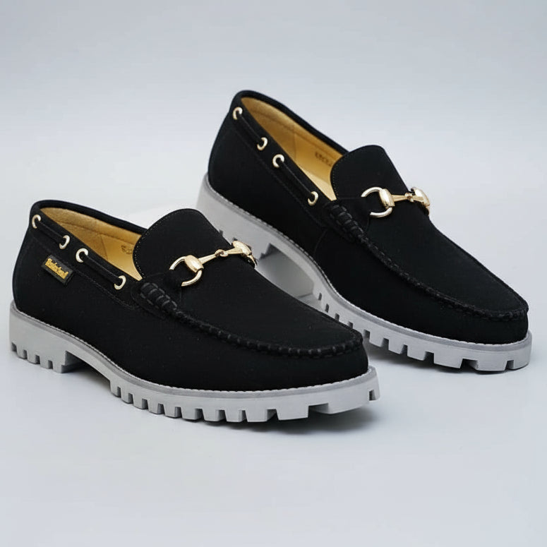 Black loafers with gold accents on a surface with a 'welcome' sign in the background