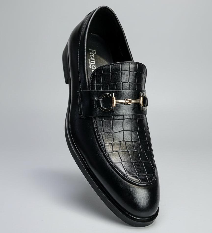 Black crocodile-patterned loafer with gold buckle held by a hand in a car interior.
