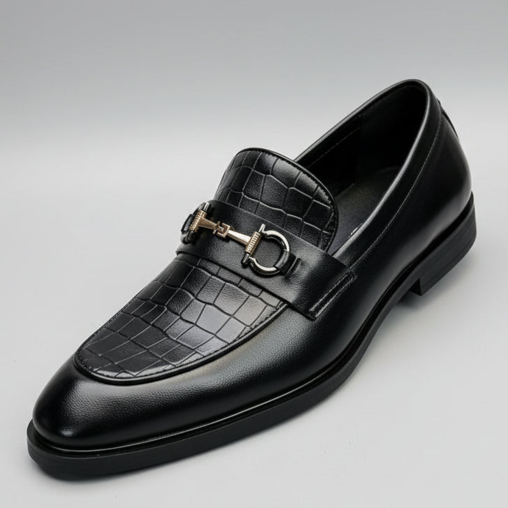 Black crocodile-patterned loafer with gold buckle held by a hand in a car interior.