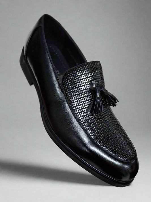 Black loafer shoe with tassel held by a hand against a neutral background